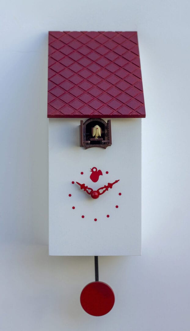 A white cuckoo clock which resembles a birdhouse, with red roof, dots for number markers, hands and pendulum