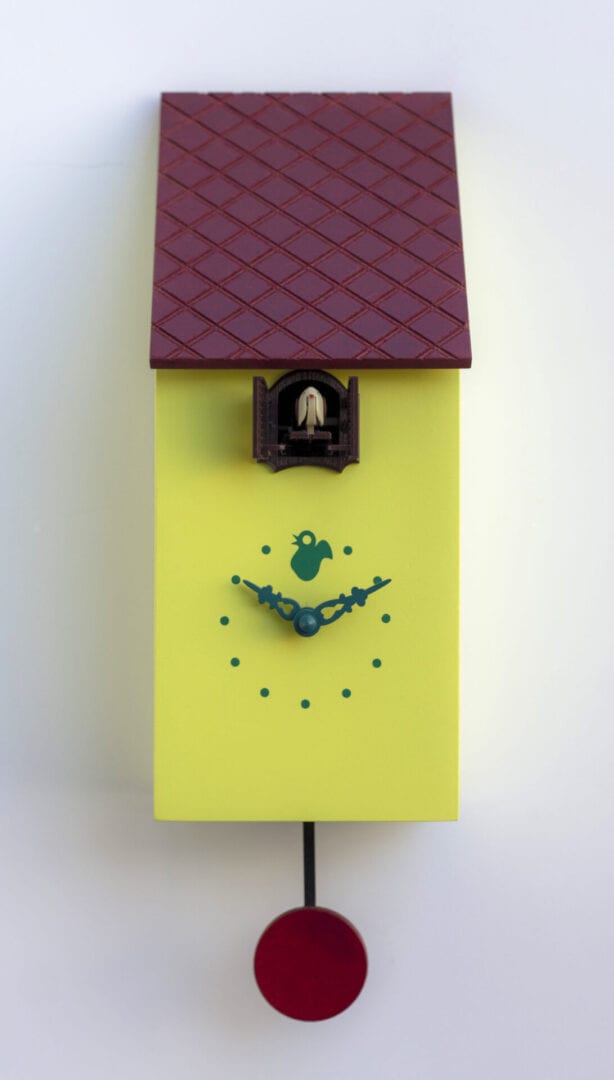 A yellow clock which looks like a bird house, with a small clockface with dotss for the numbers above which is a cuckoo house from which emerges the cuckoo. The clock has a roof which appears to be tiles and which is burgundy red