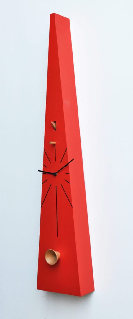 Tall cuckoo clock in red, a very narrow triangular shape, with a cuckoo house at the top and an acoustic trumpet made of beechwood at the bottom. Oversized hands add to the funky look