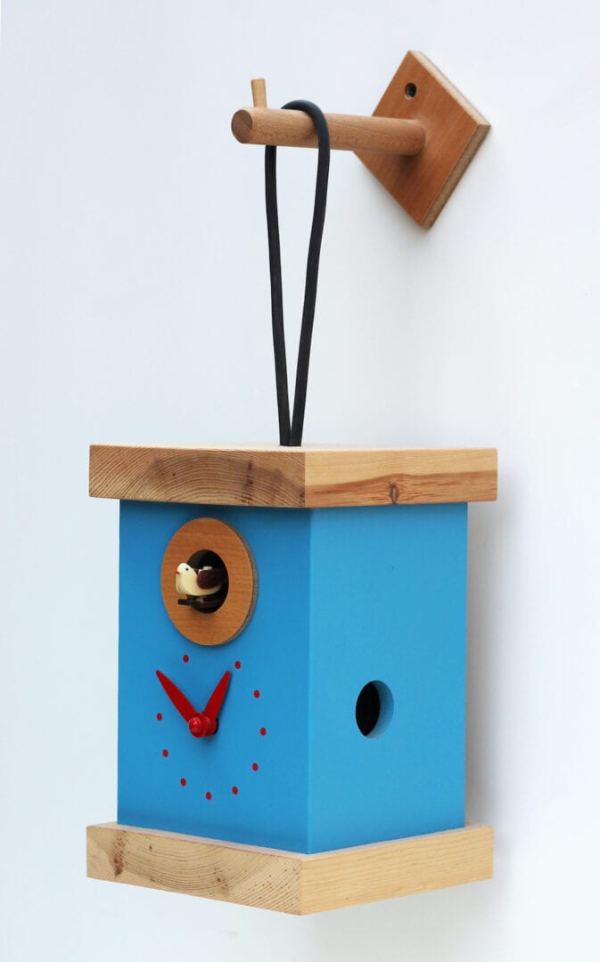 a light blue and pine coloured clock in the form of a bird box, viewed from the right hand side. The clock hangs from the wall via a hanging loop attached to a pine hook and the clockface has red detailing.