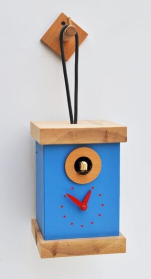A light blue square bird box shaped cuckoo clock with pine roof and base, and a hanging loop which hooks onto a pine wall hook. The detail on the clockface is red.