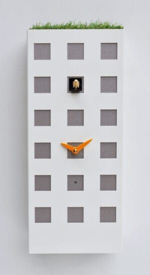An unusually styled clock in the form of a Hong Kong appartment block, in metallic white with silver coloured windows, one of which is the home for the cucckoo, and an unusual artifical grass roof.