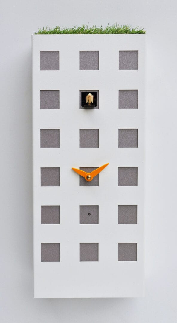 An unusually styled clock in the form of a Hong Kong appartment block, in metallic white with silver coloured windows, one of which is the home for the cucckoo, and an unusual artifical grass roof.