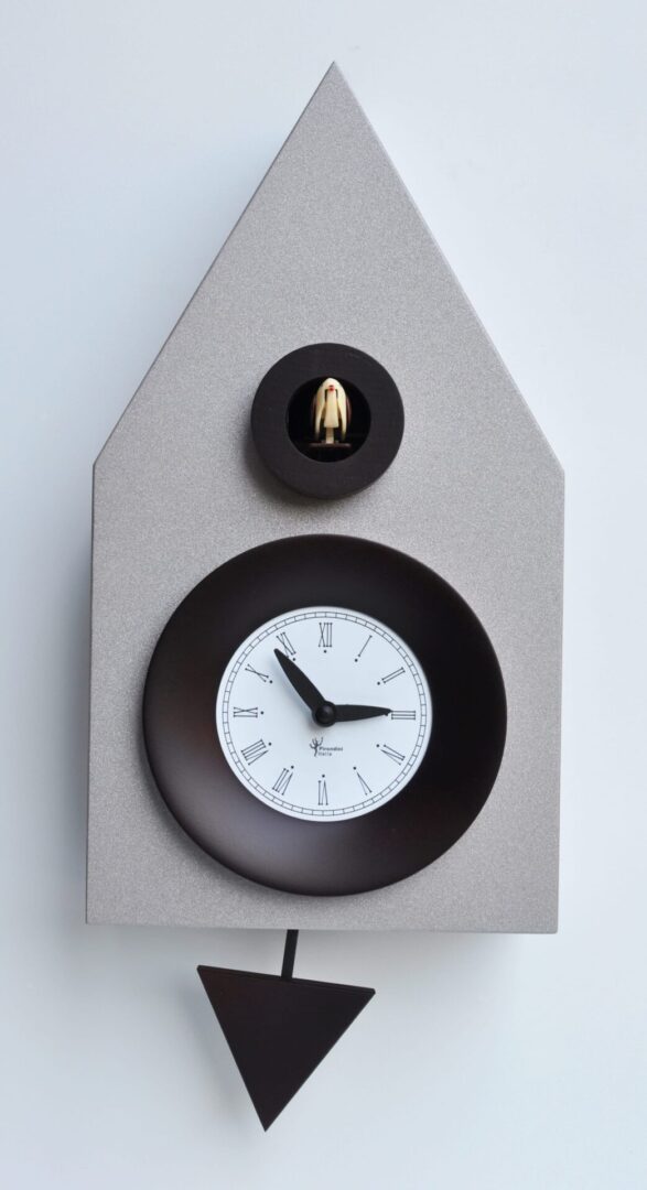 A modern take on the traditional chalet cuckoo clock, with simple lines. The center of the clock has a traditional face with bold black hands and a black surround, and the cuckoo pops out from a hole just above, with a smaller surround. the clock is in an aluminium/silver colour